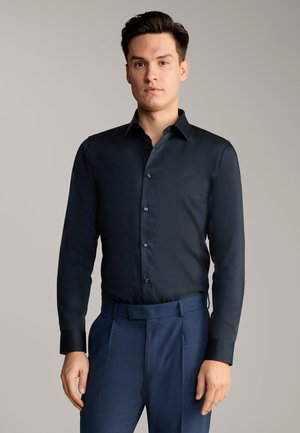 Dunkelblaues Hemd mit langen Ärmeln, glatter Textur und kontrastierenden schwarzen Manschetten. Getragen mit maßgeschneiderten blauen Hosen.