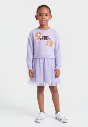 Jeune fille portant un sweat Paw Patrol lilas et une jupe en tulle assortie avec des baskets blanches, debout devant un fond clair uni.