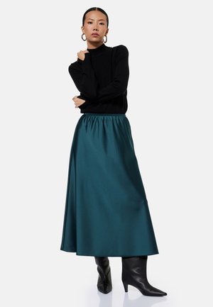 Kvinde iført en sort top med lange ærmer, teal satin midi nederdel, sorte hælede støvler og guld øreringe formet som hoops, stående med armene krydset.