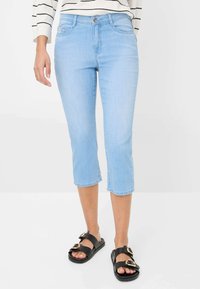 Jean cropped bleu clair en denim avec cinq poches et une fermeture à bouton, associé à des sandales noires avec des accents dorés.
