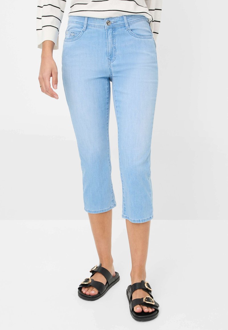 Jean cropped bleu clair en denim avec cinq poches et une fermeture à bouton, associé à des sandales noires avec des accents dorés.