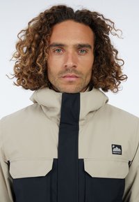 Beige und schwarze Jacke mit hohem Kragen, zwei Vordertaschen und einem Logo-Patch; glatte Textur und modernes Design.