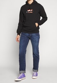 Sudadera con capucha negra con un diseño gráfico colorido en el frente, combinada con jeans azules y zapatillas moradas. Ajuste casual con capucha ajustable.