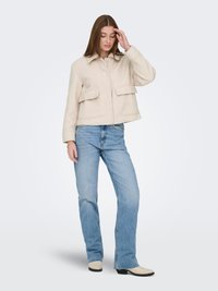 Veste matelassée couleur crème avec col, fermeture à pressions à l'avant et deux grandes poches. Associée à un jean bleu clair et des bottines beige.