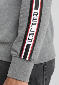 Felpa grigia con una banda a righe nere, rosse e bianche sulla manica, con il testo "REPLAY" in lettere maiuscole nere.