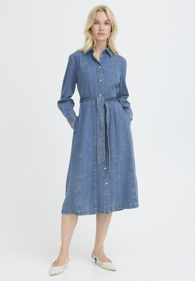 Femme blonde portant une robe en jean à manches longues boutonnée avec une ceinture à la taille et des chaussures blanches à petit talon, debout devant un fond uni.
