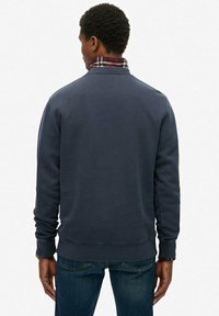 Man in een donkerblauwe sweatshirt over een geruit shirt, staande met zijn rug naar de camera tegen een effen lichte achtergrond.