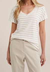 Randig V-ringad t-shirt i krämfärg och ljusbeige, tillverkad av mjukt tyg. Korta ärmar med rullade manschetter, avslappnad passform. Kombinerad med beiga byxor.