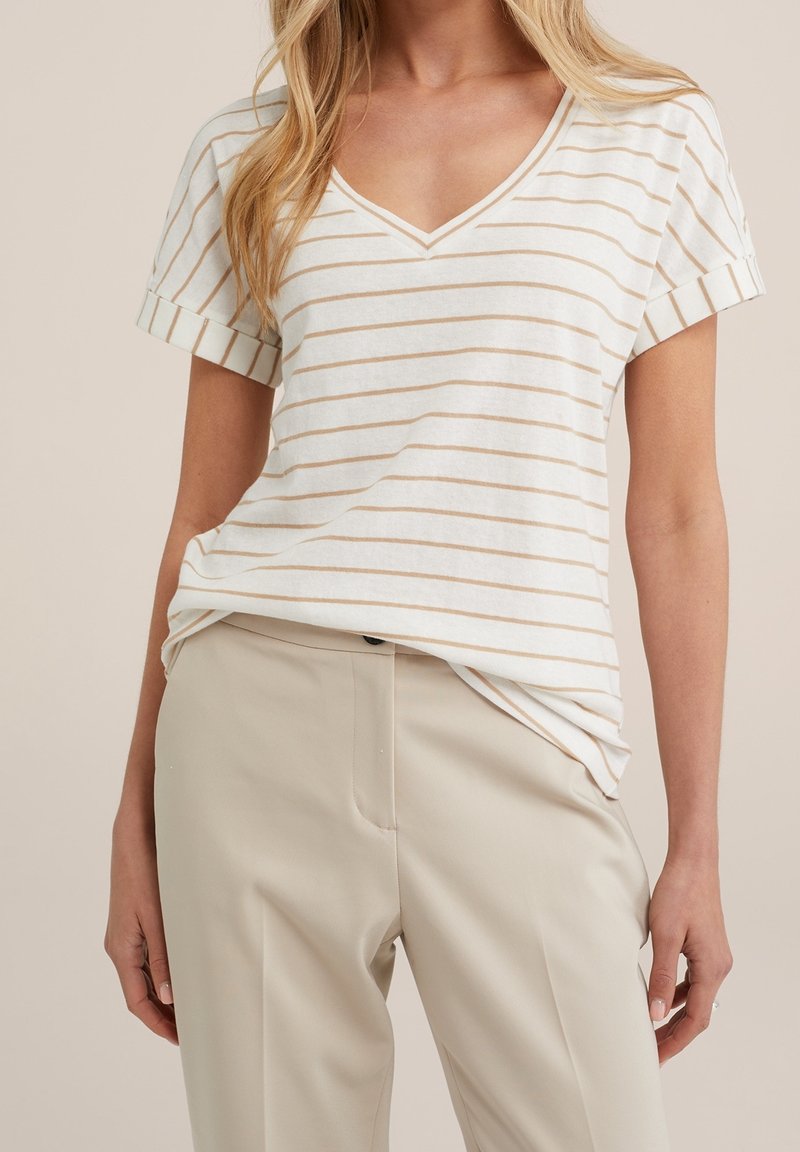 Randig V-ringad t-shirt i krämfärg och ljusbeige, tillverkad av mjukt tyg. Korta ärmar med rullade manschetter, avslappnad passform. Kombinerad med beiga byxor.