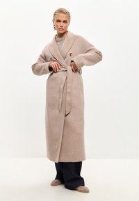 Cappotto lungo in misto lana beige con colletto a scialle, chiusura a doppiopetto, vita con cintura e tasche laterali, indossato sopra pantaloni a gamba larga di colore scuro.