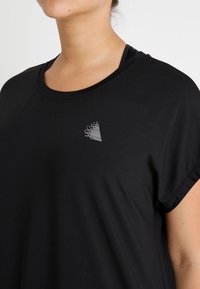 Svart atletisk t-shirt med korta ärmar, med en liten vit geometrisk logotyp på övre vänster sida. Materialet verkar glatt och lätt.