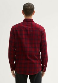 Czerwona koszula flanelowa w kratę z ciemnoniebieskimi kwadratami, z długimi rękawami, mankietami na guziki i zaokrąglonym dołem. Widok z tyłu.