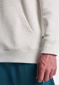 Nahaufnahme einer Hand und Taille einer Person, die einen hellbeigen Sweatshirt mit einer Taschen vorne und blauen Hosen trägt.