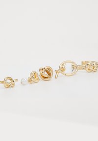 Boucles d'oreilles en métal doré présentant divers designs de nœuds, l'une ornée d'une perle blanche. Texture lisse avec finition polie et formes variées.