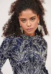 Námořnické modré šaty s propracovaným korálkovým a flitrovým květinovým vzorem, vysokým výstřihem a dlouhými rukávy. Velké náušnice vynikají a doplňují celkový design.