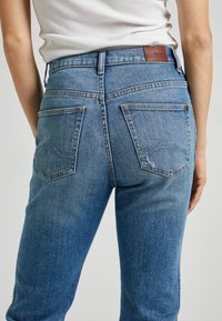 Osoba v modrých denimových kalhotách se zadními kapsami a bílém žebrovaném topu bez rukávů na jednobarevném pozadí.