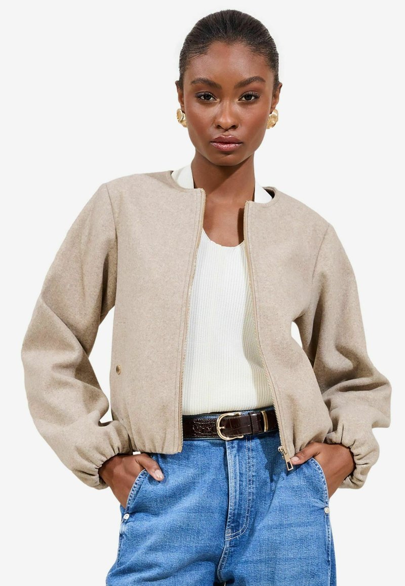 Beige zip-front jakke med rund halsudskæring, elastiske manchetter og tekstureret stof. Båret over en hvid ribbet top og blå jeans.