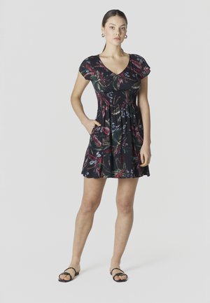 Mujer de pie con una mano en el bolsillo, vistiendo un vestido corto negro con flores y sandalias negras, frente a un fondo gris liso.