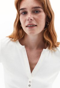 Bílý ribovaný top s kulatým výstřihem a třemi knoflíky vpředu. Modelka má vlasy stažené na stranu a minimální doplňky.