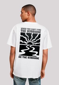 Weißes Baumwoll-T-Shirt mit einem großen schwarzen Grafikdesign auf der Rückseite, das eine Sonne und wellenförmige Linien zeigt, begleitet von Text. Kurzärmlig, lockerer Schnitt.