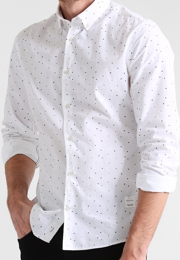 Homme portant une chemise blanche à boutons avec de petits points noirs et manches retroussées, mains partiellement dans les poches, visible du cou aux hanches.