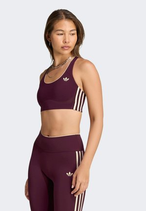 Mujer con top deportivo y leggings Adidas color granate con rayas crema, de pie y mirando hacia su izquierda sobre un fondo liso.