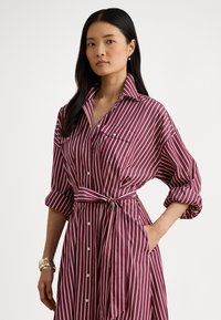 Rødbrun stripete skjortekjole med oppbrettede ermer, knapper foran, krage, midjebånd og to lapplommer. Myk stofftekstur.