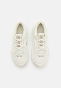 Pár smetanově bílých běžeckých bot Nike s síťovinovou texturou a hnědými logy Nike, viděné zeshora na bílém pozadí.