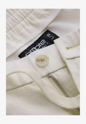 Tessuto di cotone bianco con chiusura a bottoni, dettaglio con zip e un'etichetta nera con testo bianco. Texture liscia con una leggera lucentezza.