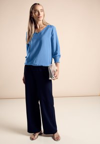Blusa blu, ampia e morbida, con maniche a tre quarti e scollatura a V. Abbinata a pantaloni navy a gamba larga e una pochette metallizzata argentea.