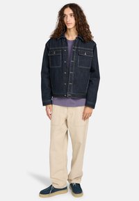Giubbotto in denim scuro con chiusura a bottoni, due tasche sul petto e cuciture a contrasto. Abbinato a una camicia viola e pantaloni beige.