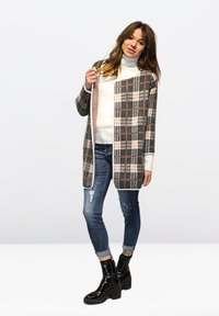 Cardigan a cuadros en gris, rosa y blanco roto, con diseño de frente abierto, combinado con un suéter de cuello alto blanco, y pantalones vaqueros azules desgastados y botas negras.
