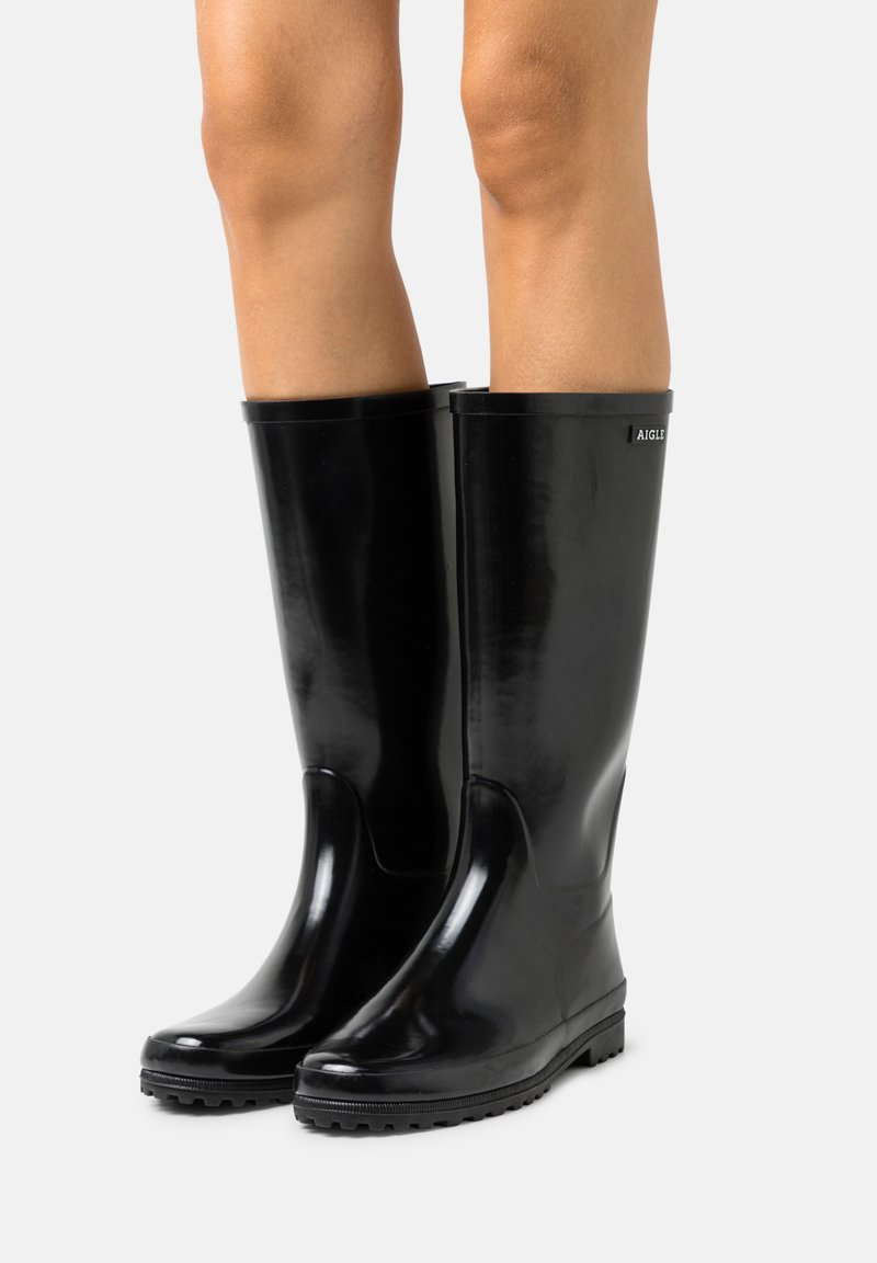 Bottes de pluie en caoutchouc noires, arrivant au genou, avec une texture lisse, un bout rond et une semelle antidérapante. Présente un accent de logo près du haut.