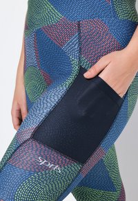 Pantylegging met een kleurrijk, abstract patroon in blauw, groen, rood en marineblauw, met een mesh zak aan de zijkant en een logo op het onderbeen.