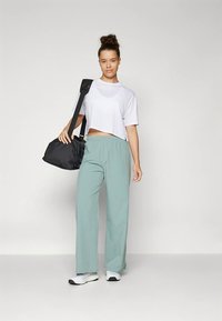 Jauna moteris stovi su juodu sportiniu maišu ant peties, apsirengusi balta trumpine marškinėliais, šviesiai žaliais plačiais džinsais ir baltais sportbačiais.