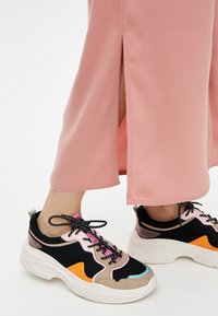 Des baskets épaisses composées d'un mélange de matériaux noirs, roses, oranges et bleus, avec des superpositions texturées et des lacets noirs. Portées avec une longue jupe rose.