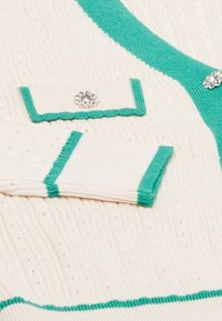 Krämfärgad stickad cardigan med turkos kant. Innehåller dekorativ hårdvara och textil, med betoning på ribbade och släta sektioner.