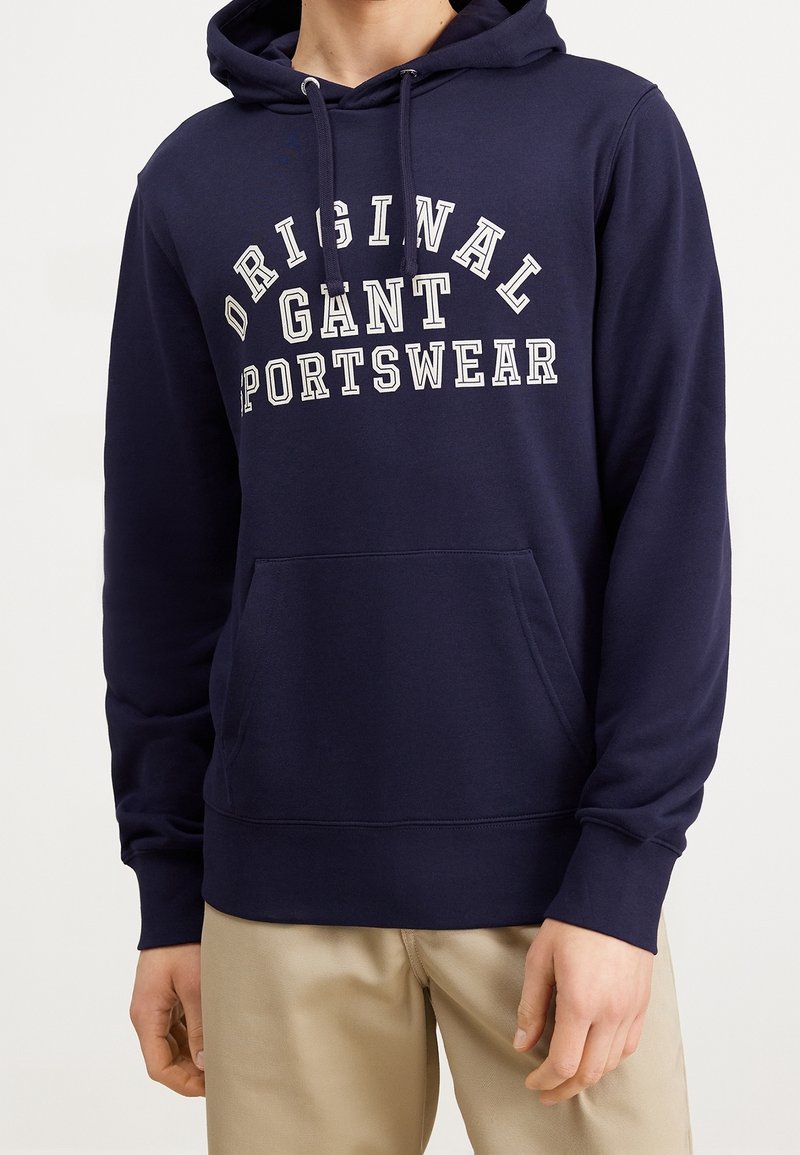 Sweat à capuche bleu marine avec texte blanc "ORIGINAL GANT SPORTSWEAR", poche kangourou, cordons, porté avec un pantalon beige, torse et bras visibles.