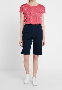 Camiseta de manga corta rosa con estampado floral, combinada con shorts azul marino de longitud a la rodilla y zapatillas blancas. Diseño simple, ajuste casual.