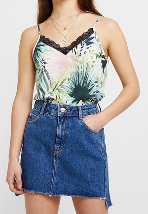 Kvinde iført en ærmeløs blomsterprintet top med sort blondekant og en blå denimnederdel med forlommer, den ene hånd i lommen, mod en ensfarvet baggrund.
