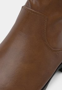 Gros plan sur une botte à cheville en cuir brun lisse avec une couture visible et un bord de semelle noir.