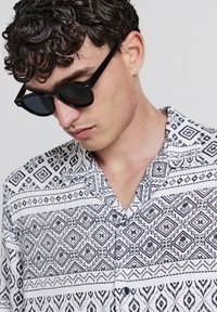 Chemise à manches courtes noir et blanc avec des motifs géométriques, tissu tissé, et col. Accompagnée de lunettes de soleil foncées.