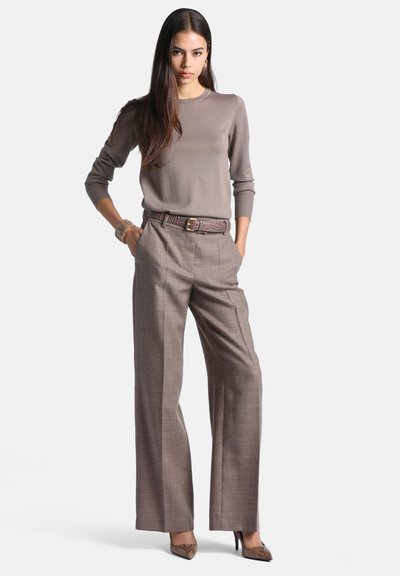 Femme aux longs cheveux foncés, portant un pull taupe, un pantalon marron taille haute et jambes larges, et des talons hauts à motif peau de serpent, mains dans les poches.
