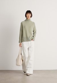 Woman walking indoors wearing a light green turtleneck sweater, white wide-leg pants, beige shoes, and carrying a large beige tote bag.