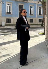 Frau im schwarzen Anzug und Sonnenbrille hält eine schwarze Clutch und steht auf dem Bürgersteig der Stadt vor einem blauen Gebäude mit weißen Fenstern.