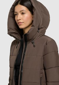 Braune Daunenjacke mit großer Kapuze, mit Reißverschluss, elastischen Kordelzügen und gestepptem Muster. Über einem schwarzen Oberteil getragen.