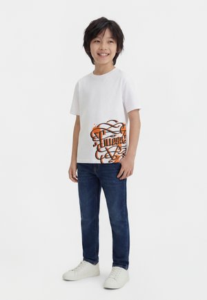 Boy smiling, wearing white T-shirt with orange graffiti design, blue jeans, and white sneakers, standing against plain white background.