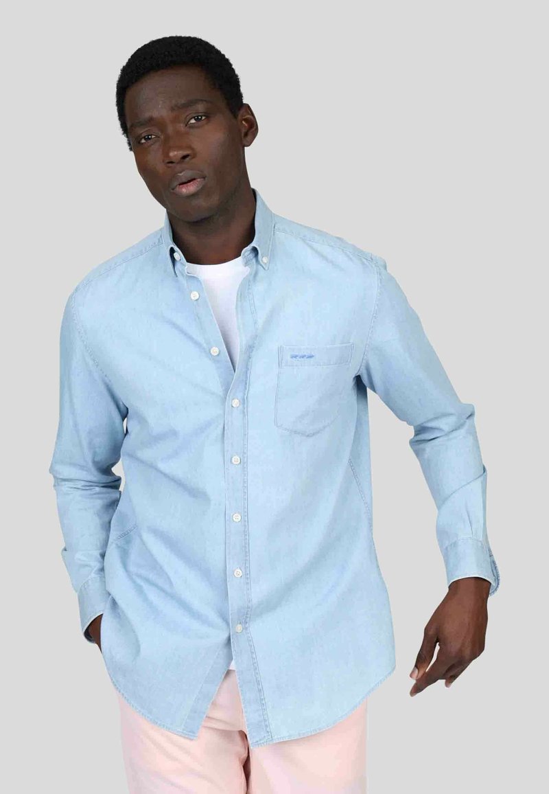 Chemise en denim bleu clair avec un col boutonné, une poche poitrine simple et des manches longues, portée par-dessus un t-shirt blanc.