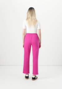 Kvinne med langt blondt hår, iført en hvit topp, lyserosa bukser og svart-hvite joggesko, stående mot en ensfarget hvit bakgrunn.