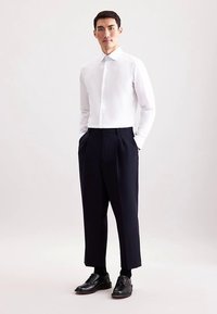 Witte blouse met lange mouwen en een rechte kraag, gecombineerd met marineblauwe geplooide broek en zwarte leren schoenen, voor een formele uitstraling.